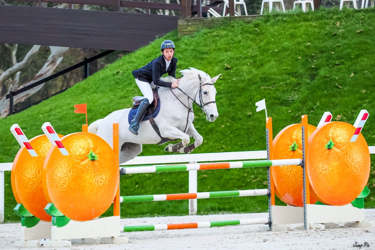 Maxence Violleau et sa jument blanche Confidence lors d'un concours de saut d'obstacles. Ils franchissent un oxer encadré de deux chandeliers en forme d'orange.