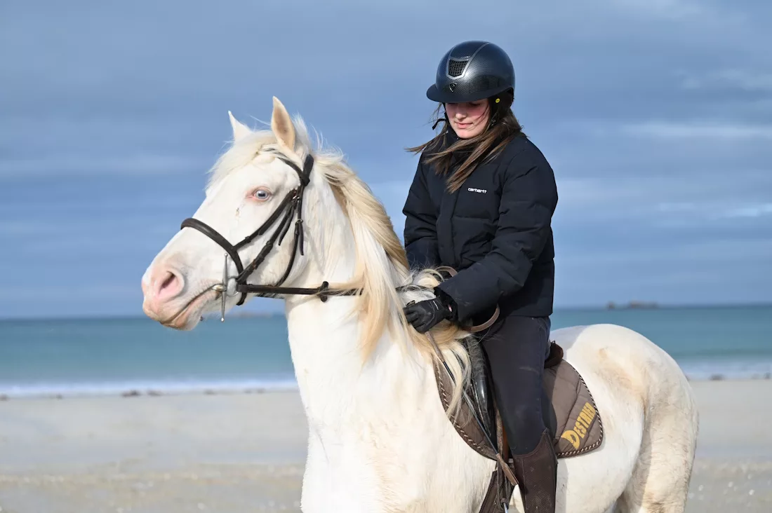 poney de club blanc monté par une jeuen cavalière lors d'unes ortie à la plage en Finsitère